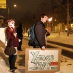 EIn Mann hälz neben einer Haltestelle ein Schild in der Hand auf dem "X-MAS Tram Check In" zu lesenist. Neben ihm steht eine Frau mit Engelsflügeln